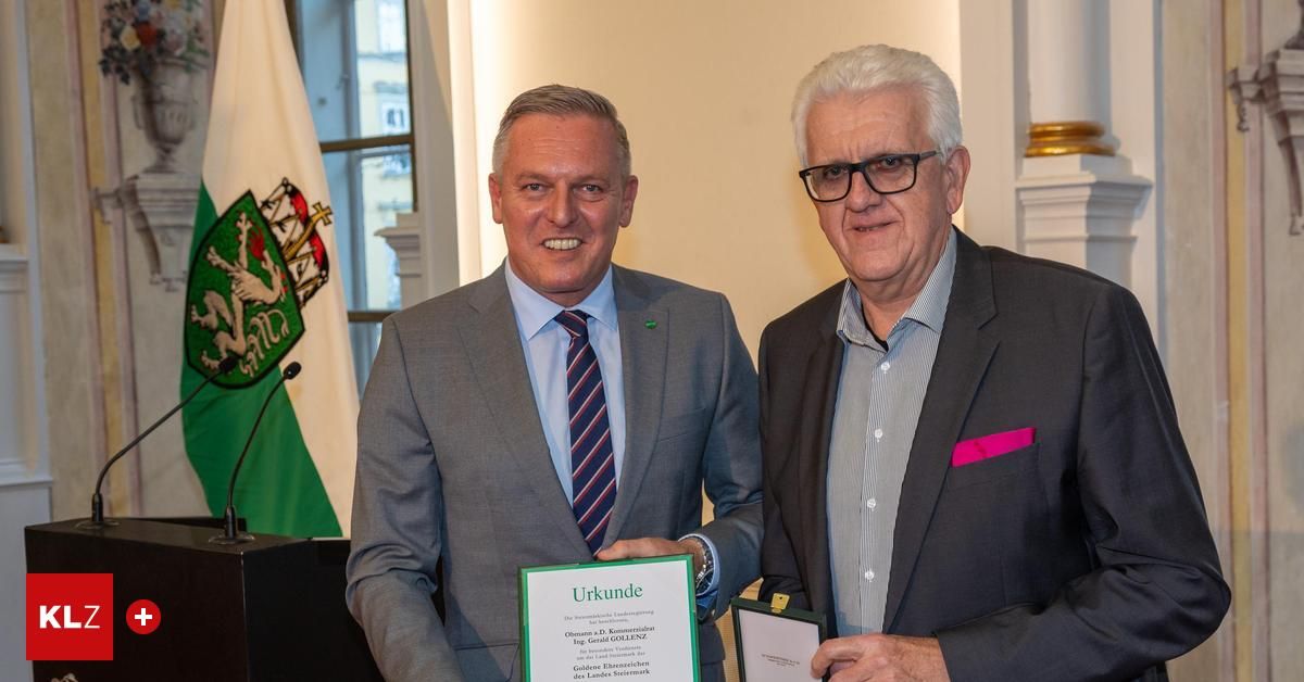 Zwei Männer stehen lächelnd nebeneinander. Sie halten ein gerahmtes Zertifikat und eine Schachtel. Hinter ihnen ist eine Flagge mit einem Wappen und ein Podium mit einem Mikrofon.