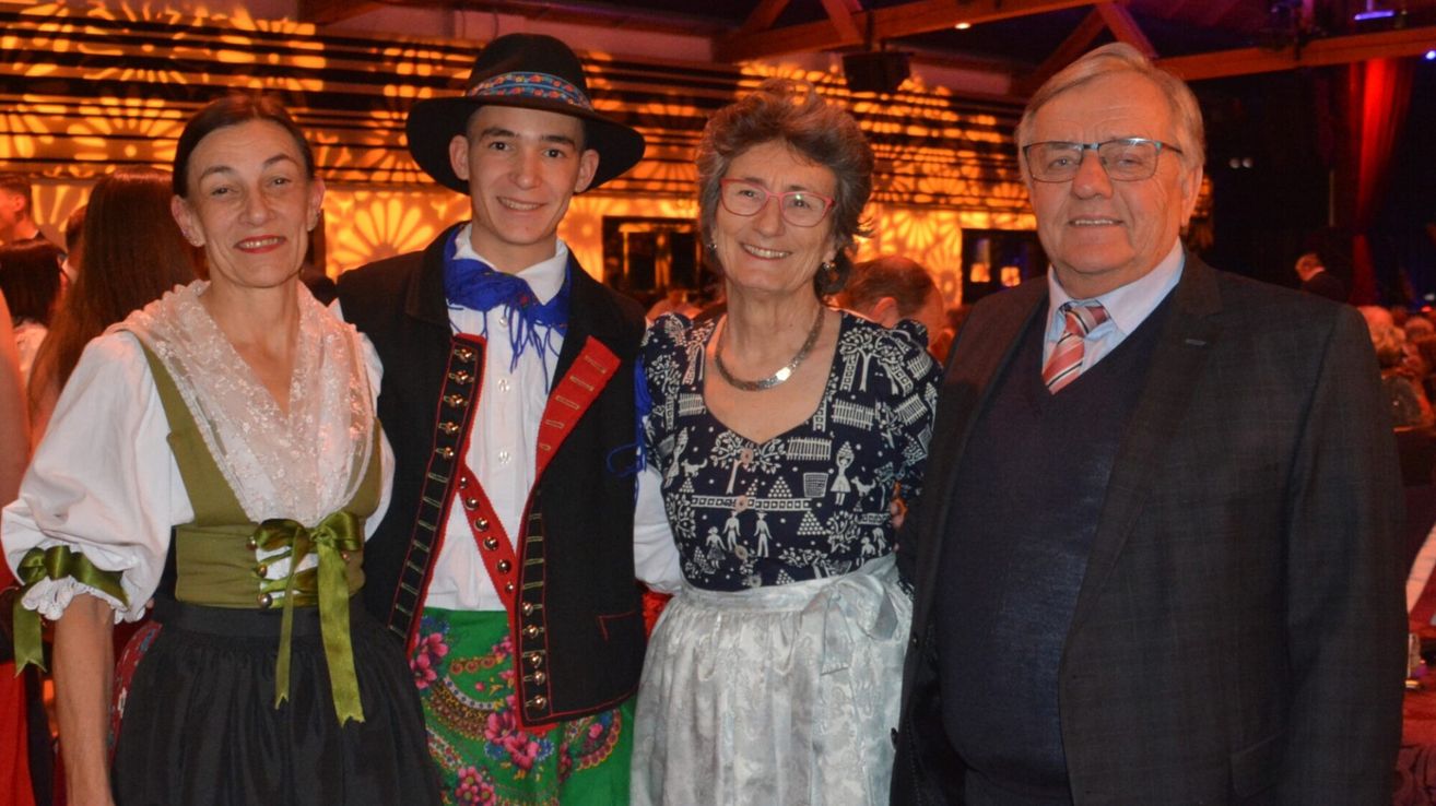 Drei Personen, eine in traditioneller Kleidung, lächeln für ein Foto bei einer Veranstaltung. Hinter ihnen sind weitere Personen anwesend, und der Hintergrund ist schwach beleuchtet.