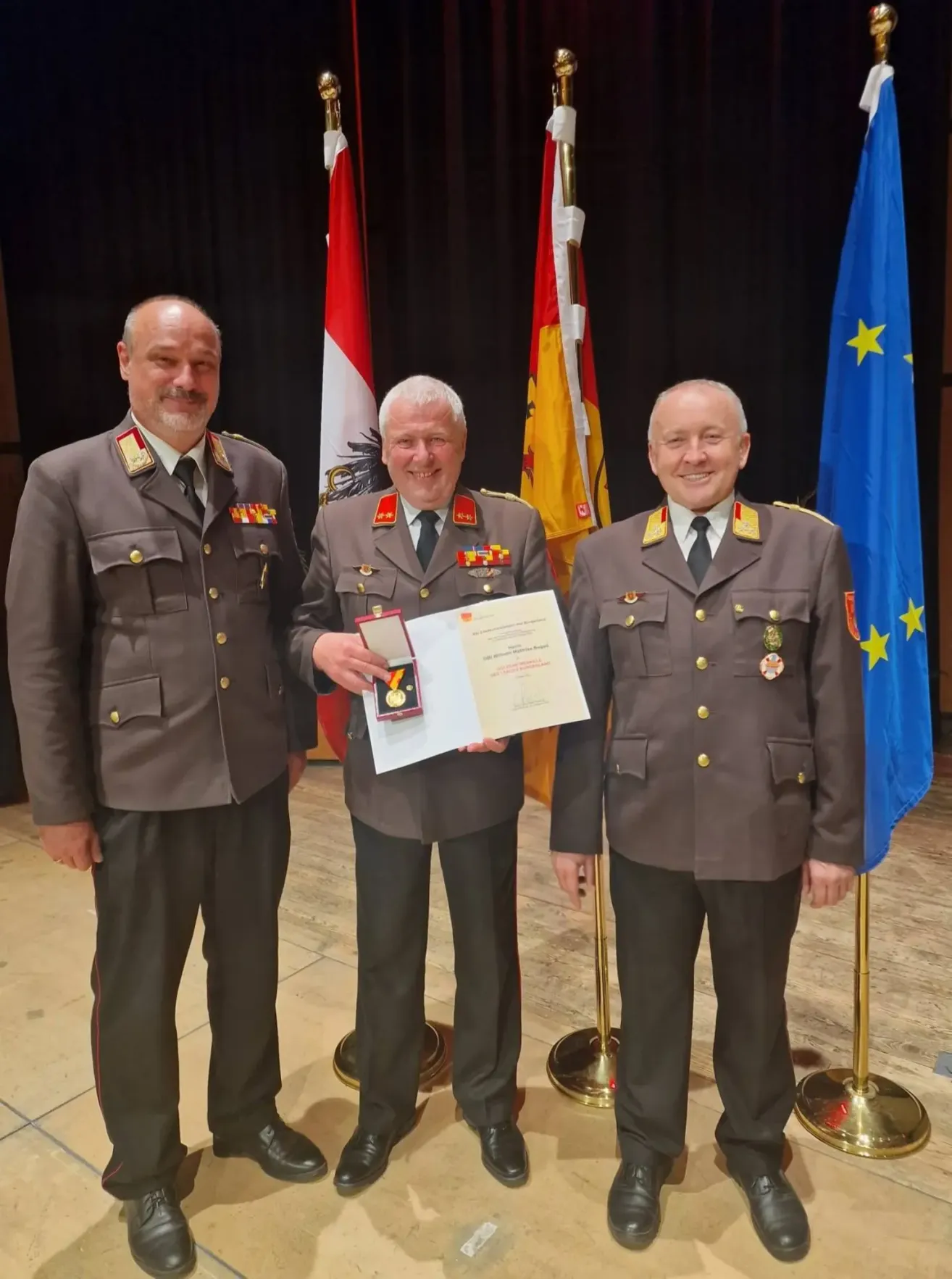 Drei Männer in Militäruniformen stehen nebeneinander, einer hält ein Zertifikat und eine Medaille und lächelt für ein Foto mit Flaggen im Hintergrund.