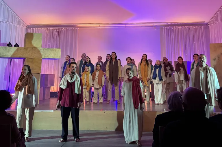 Ein Chor tritt mit einem Kreuz im Hintergrund auf der Bühne auf, beleuchtet von lila Lichtern und in traditioneller Kleidung.