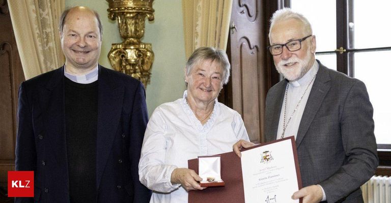 Eine Frau in einem weißen Hemd lächelt, während sie ein Diplom und eine Medaille hält, flankiert von zwei Männern in formeller Kleidung, möglicherweise während einer Preisverleihung.