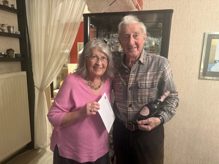 Ein älteres Paar steht eng beieinander, hält ein Papier und eine Flasche Wein in der Hand, lächelt und schaut in die Kamera in einem Raum mit einem Vorhang und einem Schrank.