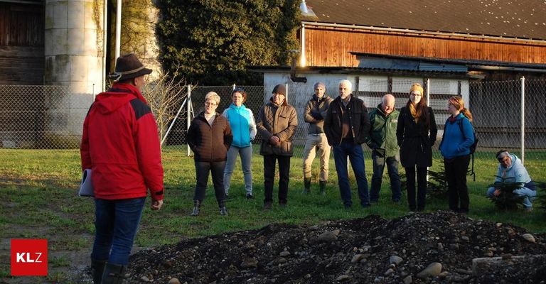 Eine Gruppe von Erwachsenen steht auf Gras in der Nähe eines Bauernhauses, mit einem Haufen Steine im Vordergrund. Einige tragen Mützen und Stiefel. Eine Frau in einer blauen Jacke schaut etwas an, während andere aufmerksam sind.