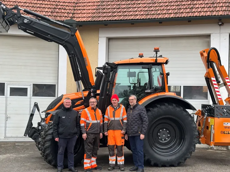 Vier Männer stehen vor einem großen orangefarbenen Traktor. Dahinter befindet sich ein Gebäude mit einem roten Dach. Der Mann links trägt eine Brille und einen schwarzen Mantel.