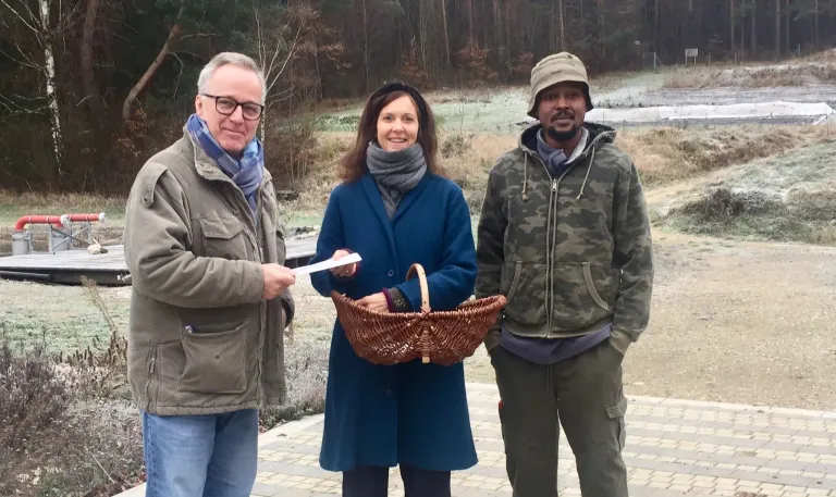 Drei Personen stehen vor einem Wald. Ein Mann auf der linken Seite hält ein Papier, eine Frau hält einen Korb und ein Mann auf der rechten Seite lächelt.
