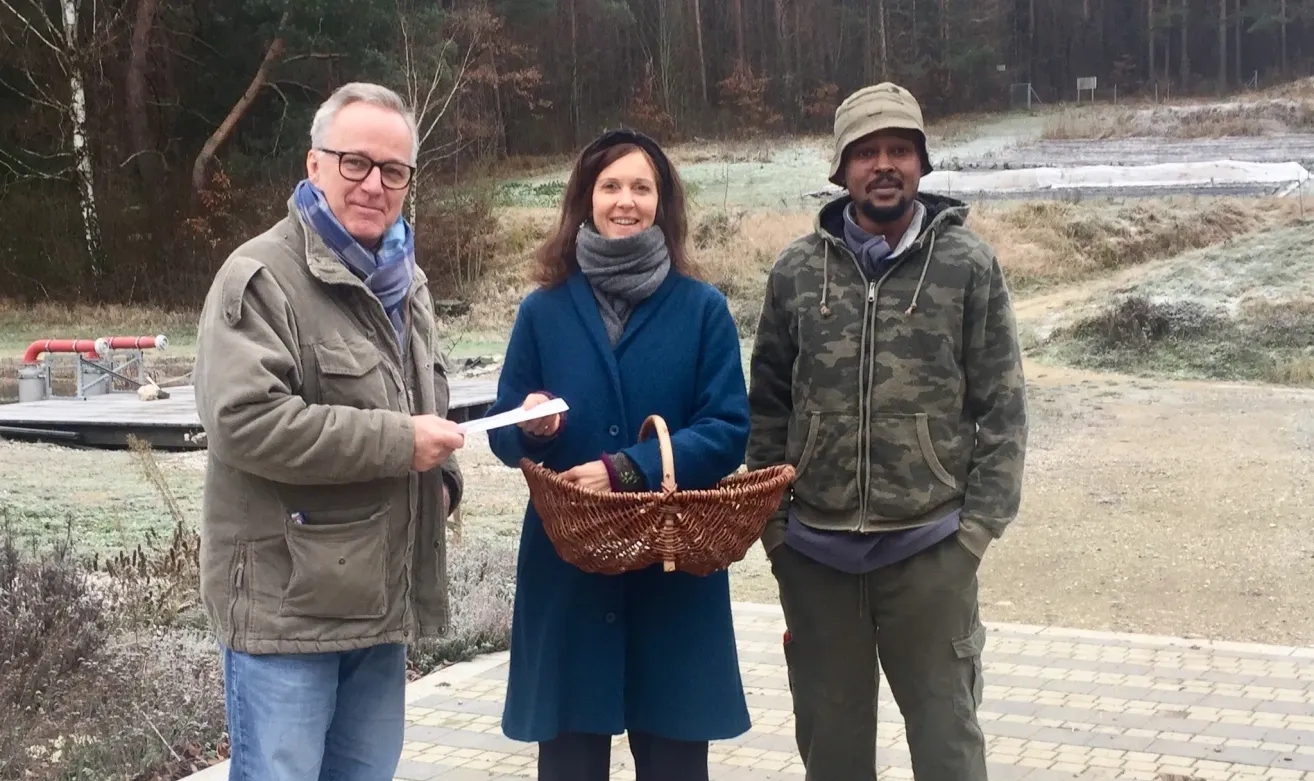Drei Personen stehen vor einem Wald. Ein Mann auf der linken Seite hält ein Papier, eine Frau hält einen Korb und ein Mann auf der rechten Seite lächelt.