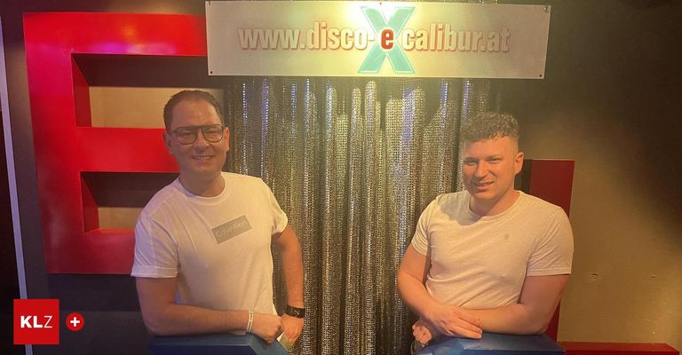 Zwei Männer stehen lächelnd vor einem Disco-Hintergrund mit einem Schild, das 'www.disco-e-calibur.at' steht. Beide tragen weiße Calvin Klein T-Shirts. Der auf der linken Seite hat eine Uhr und Armbänder.
