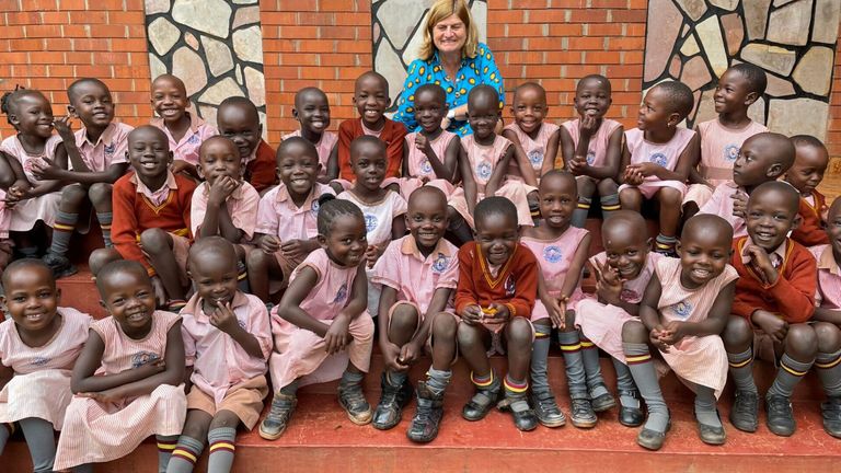 Eine Frau in einem blauen Shirt sitzt hinter einer Gruppe lächelnder Kinder in pinken und orangefarbenen Uniformen. Die Kinder sitzen auf einer Stufe mit Ziegelwänden im Hintergrund.