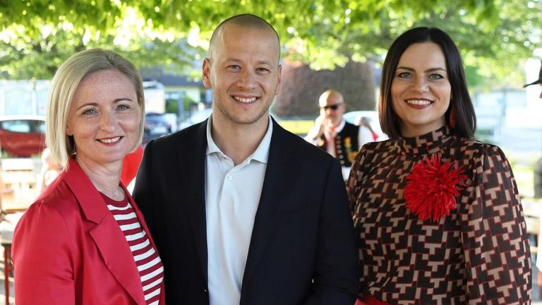 Ein Mann und zwei Frauen lächeln für ein Foto in einem Park. Der Mann trägt einen dunklen Anzug und ein weißes Hemd. Die Frauen tragen ein rotes gestreiftes Shirt und eine gemusterte Bluse. Bäume und Autos sind im Hintergrund.