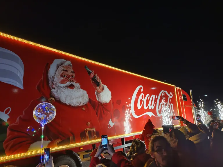 Ein großer Coca-Cola-Lkw mit dem Weihnachtsmann, der eine Flasche Coca-Cola hält. Viele Menschen sind um den Lkw herum und machen Fotos mit ihren Handys.