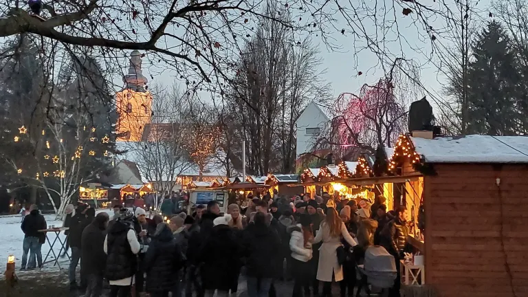 Ein überfüllter Außenbereich mit vielen Menschen, Winterdekorationen und beleuchteten Ständen. Bäume ohne Blätter und ein Gebäude mit Turm im Hintergrund.