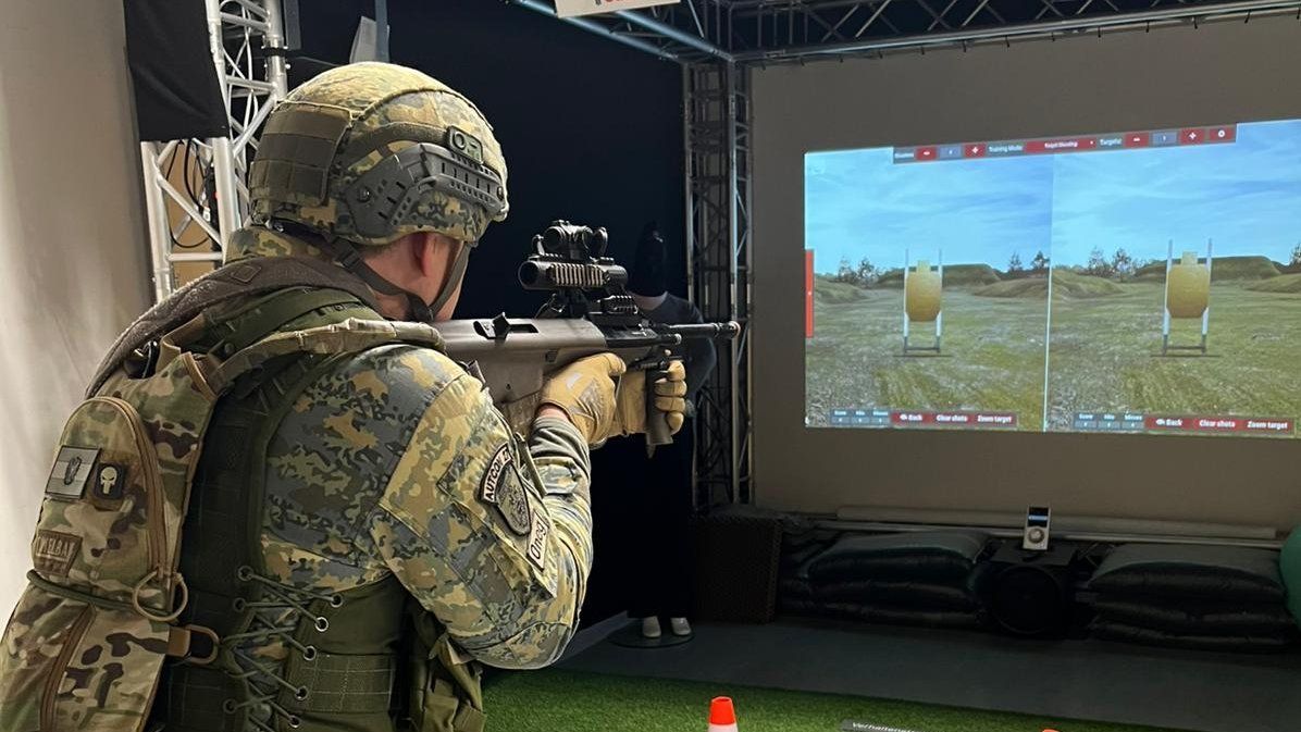 Ein Soldat in Tarnkleidung zielt mit einem Gewehr auf ein projiziertes Ziel auf einem Bildschirm.