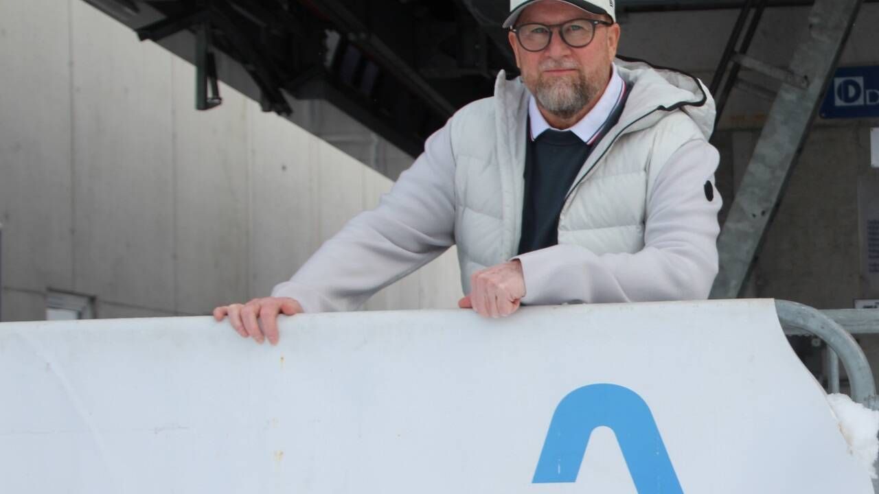 Ein Mann mit Brille und Mütze lehnt sich an eine weiße Wand mit einem blauen 'A'-Schild. Er ist in einer weißen Jacke, einem schwarzen Innenhemd und einer Krawatte gekleidet.