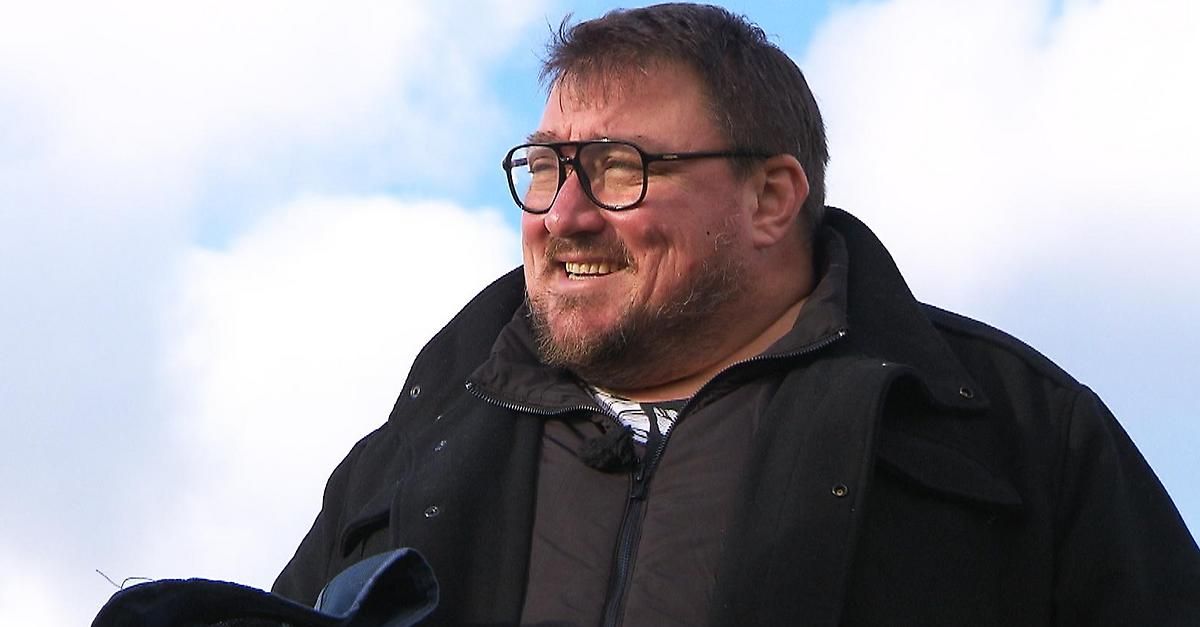 Ein Mann mit Bart und Schnurrbart lächelt und trägt eine Brille. Er ist in einen schwarzen Mantel gekleidet und ist im Freien unter einem bewölkten Himmel.