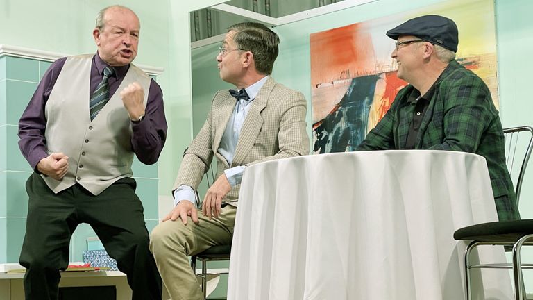 Drei Männer sitzen um einen Tisch herum. Der Mann auf der linken Seite trägt ein lila Hemd und eine Weste. Der Mann in der Mitte trägt einen Anzug mit einer Fliege. Der Mann auf der rechten Seite trägt eine grüne Jacke und einen Hut. Ein abstraktes Gemälde hängt an der Wand hinter ihnen.