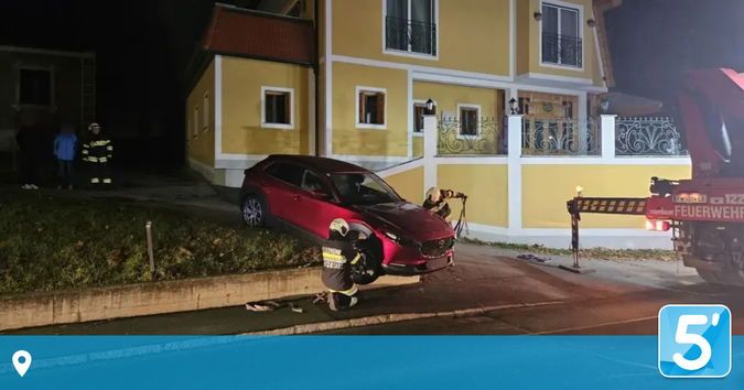 Feuerwehrleute arbeiten an einem roten Auto, das auf der Straße vor einem gelben Haus geparkt ist. Ein Feuerwehrwagen ist in der Nähe, und zwei Personen arbeiten am Auto.