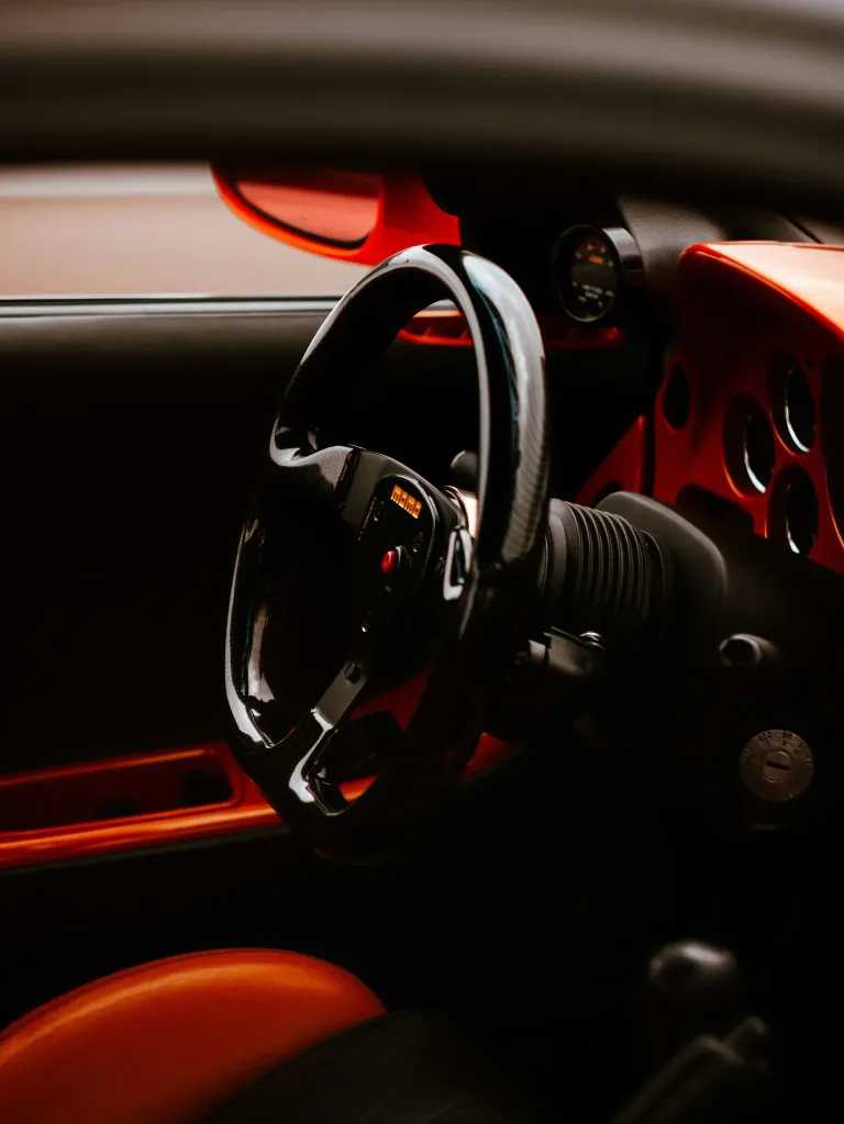 Nahaufnahme des Innenraums eines Autos mit einem schwarzen Lenkrad und rotem Armaturenbrett. Das Armaturenbrett hat verschiedene Anzeigen und Knöpfe.