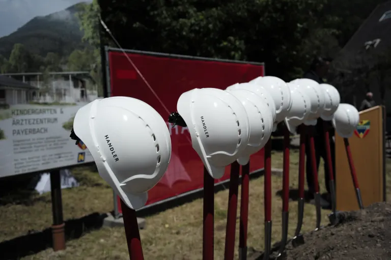 Eine Reihe weißer Bauhelme mit der Aufschrift 'HANDLER' wird auf roten Stangen in einem Grasbereich ausgestellt, mit einem roten Banner und Bäumen im Hintergrund.