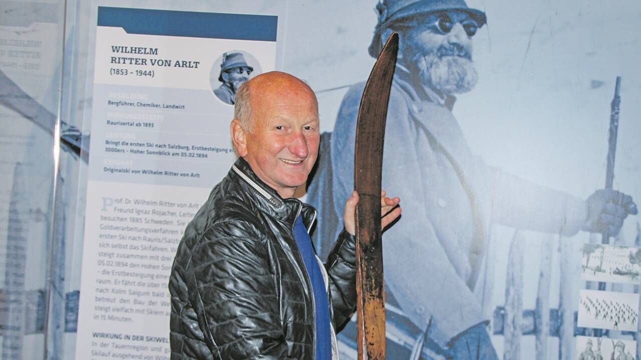 Ein älterer Mann mit Lächeln hält einen hölzernen Ski und steht vor einem Plakat eines Mannes in Uniform.