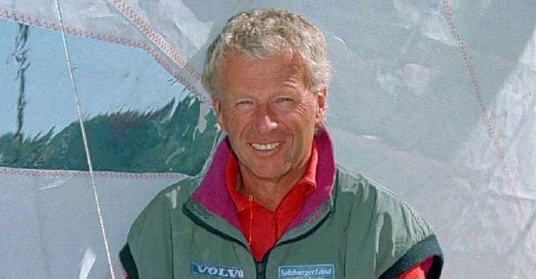 Ein Mann in einer grünen Jacke mit rotem Kragen steht vor einem verschneiten Hintergrund. Er lächelt und scheint für ein Foto zu posieren.