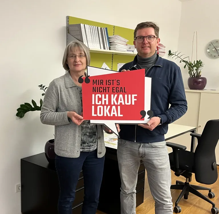 Ein Mann und eine Frau stehen in einem Büro und halten ein Schild mit der Aufschrift 'MIR IST'S NICHT EGAL ICH KAUF LOKAL'. Sie lächeln.