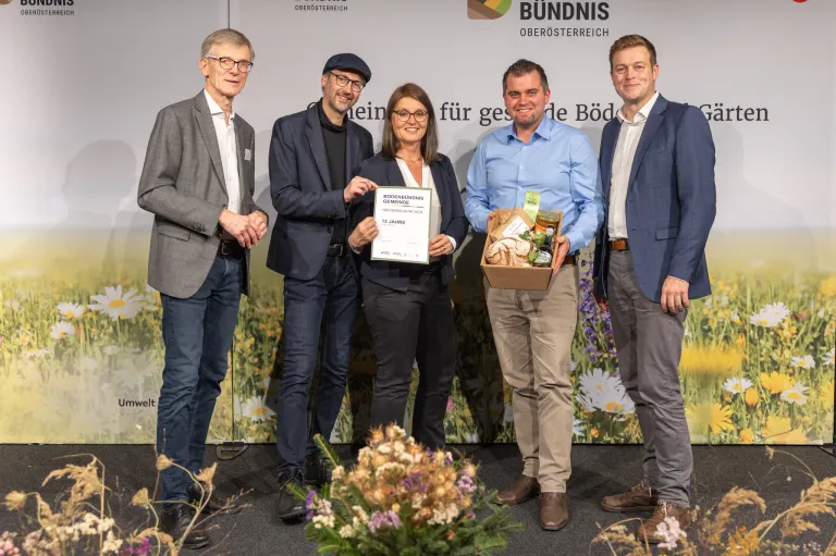 Fünf Personen stehen auf einer Bühne und lächeln, vor einem Hintergrund aus Blumen und Text. Die zentrale Figur hält ein Zertifikat und eine Box. Die beiden Männer an den Enden tragen Anzüge, während die anderen lässige Kleidung tragen.