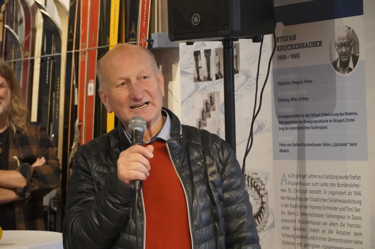 Ein Mann steht in einem Skigeschäft und hält ein Mikrofon. Hinter ihm befindet sich eine Wand mit Fotos und einer Texttafel.