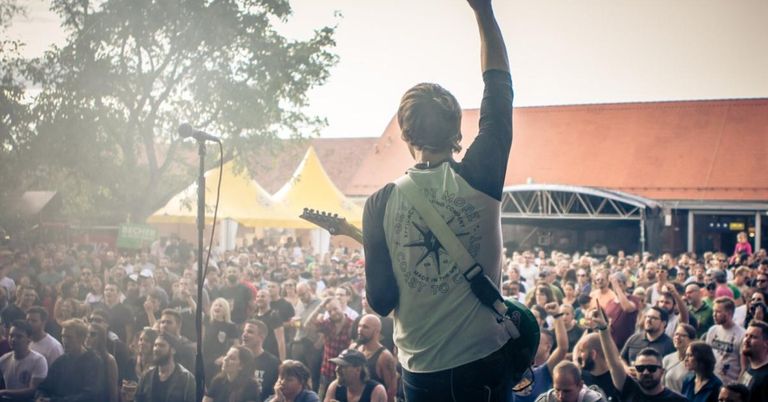 Ein Musiker mit einer Gitarre auf der Bühne, der vor einem Publikum bei einem Open-Air-Festival auftritt, mit Zelten im Hintergrund.