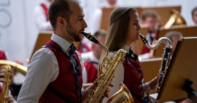 Ein Mann und eine Frau spielen Saxophon in einem Konzertsaal, beide tragen rote Westen und Fliegen. Dahinter sitzen mehrere Personen, möglicherweise andere Musiker.