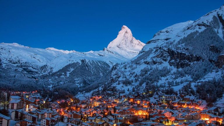 Ein verschneites Dorf am Fuße des Matterhorns unter einem klaren Nachthimmel. Das Dorf ist mit vielen Lichtern beleuchtet, während die Berge schneebedeckt sind.