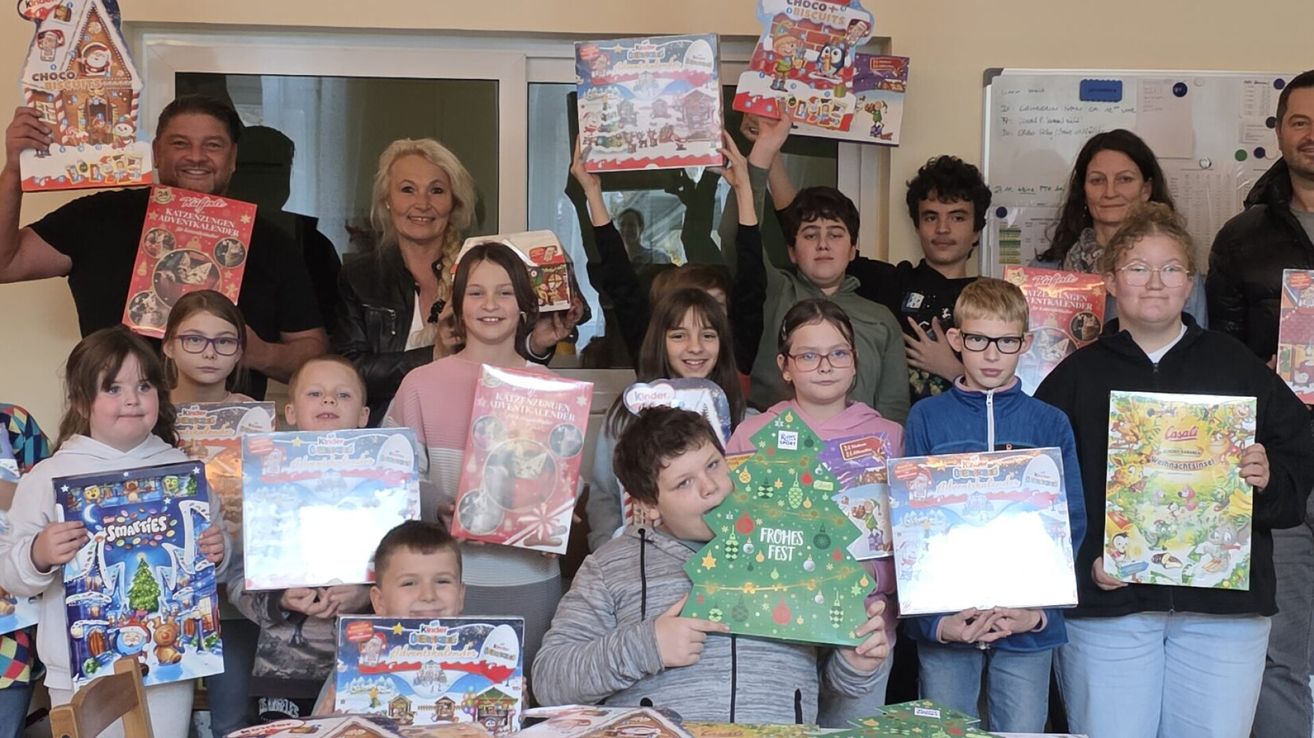 Eine Gruppe von Kindern und Erwachsenen posiert mit weihnachtlichen Geschenken. Die Kinder halten verschiedene Schachteln und Karten hoch, während eine Frau lächelt und hinter ihnen steht.