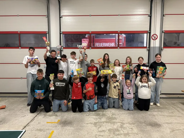 Eine Gruppe junger Menschen in einer Garage posiert mit Trophäen und Snacks. Im Hintergrund ist ein Feuerwehrauto zu sehen.