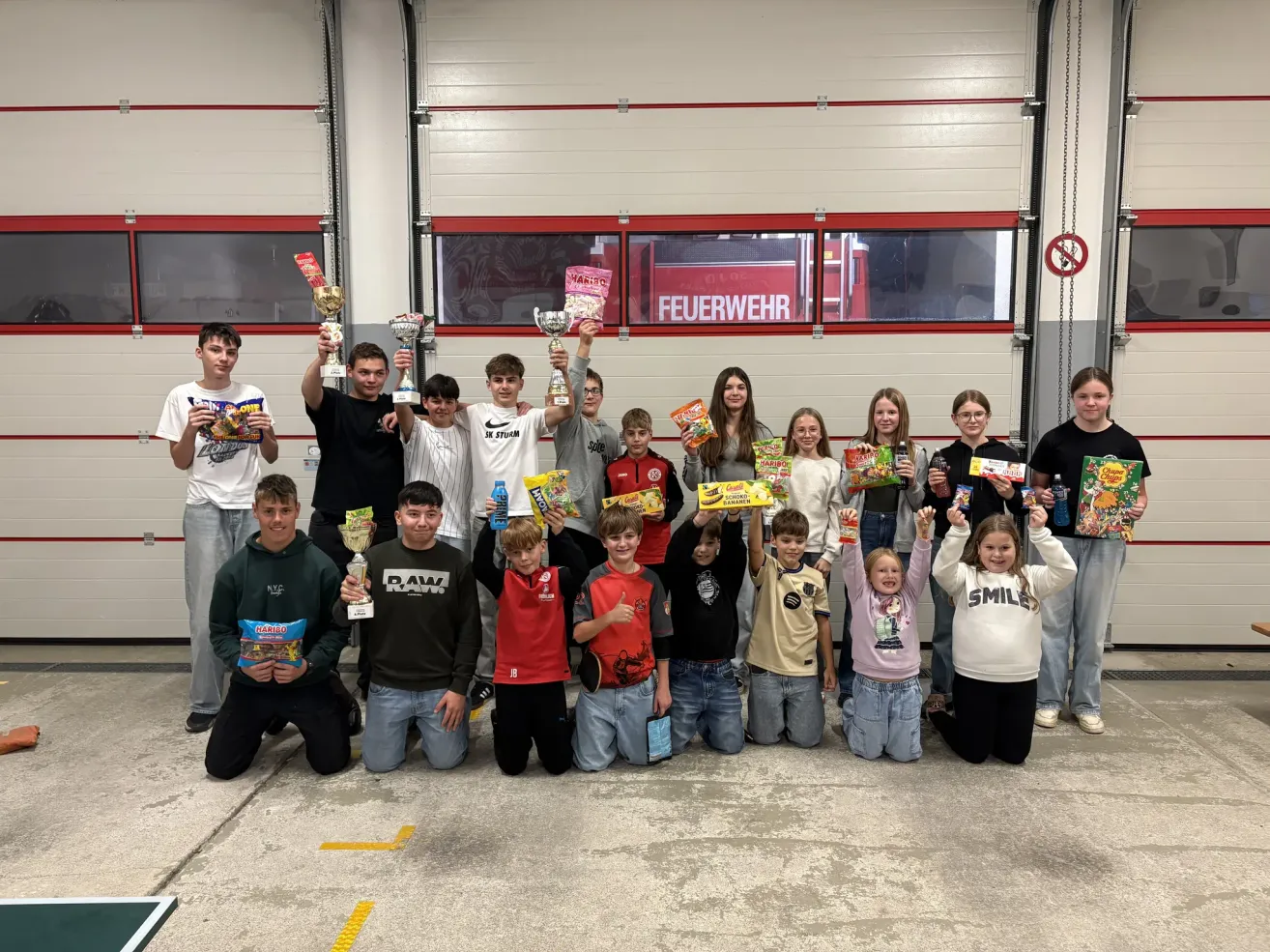 Eine Gruppe junger Menschen in einer Garage posiert mit Trophäen und Snacks. Im Hintergrund ist ein Feuerwehrauto zu sehen.
