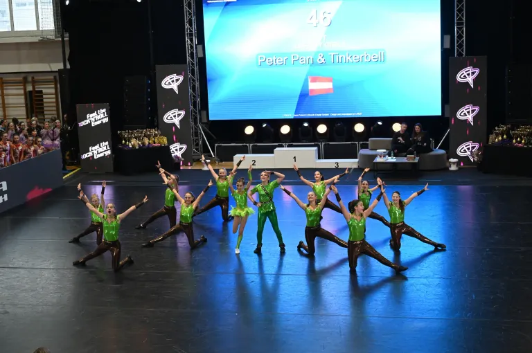 Eine Gruppe junger Frauen in grünen Kostümen tritt auf einer Bühne auf. Vor ihnen steht ein Podium mit Nummern. Eine große Leinwand zeigt die Wörter Peter Pan und Tinkerbell.