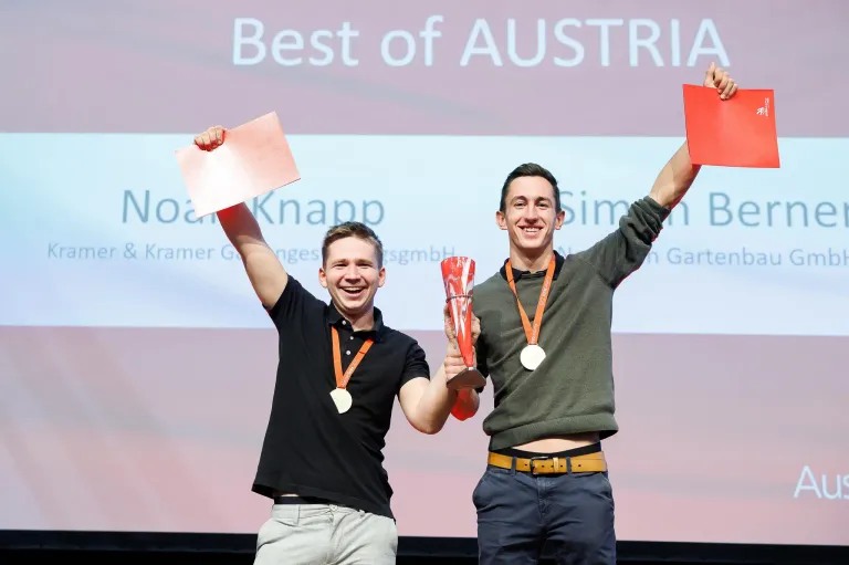 Zwei Männer feiern auf der Bühne mit Medaillen und Trophäen, ein Hintergrund sagt 'Best of AUSTRIA'.