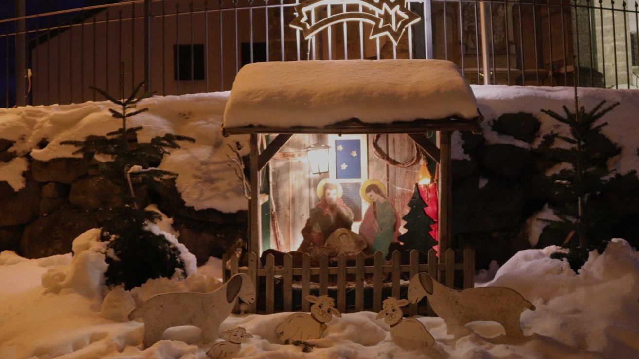 A Christmas nativity scene is displayed outdoors in the snow with a star above. The scene includes figures of Mary, Joseph, and baby Jesus. It is surrounded by snow and Christmas decorations.