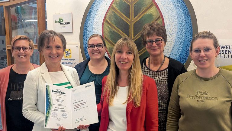 Vier Frauen stehen zusammen und halten ein Zertifikat; die Frau auf der linken Seite hält ein Zertifikat. Sie alle lächeln und schauen in die Kamera. Hinter ihnen befindet sich eine blattförmige Dekoration mit einem Schild, das 'Tut gut!' besagt.