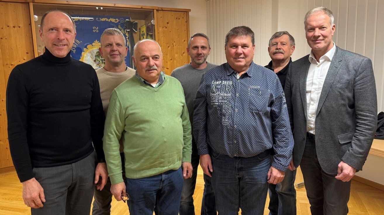 Sechs Männer stehen zusammen in einem Raum und lächeln für ein Foto. Zwei tragen lange Ärmel, einer in grün, der andere in blau mit weißen Punkten. Das blaue Hemd hat Text. Dahinter ist eine Holzwand mit einem Banner.