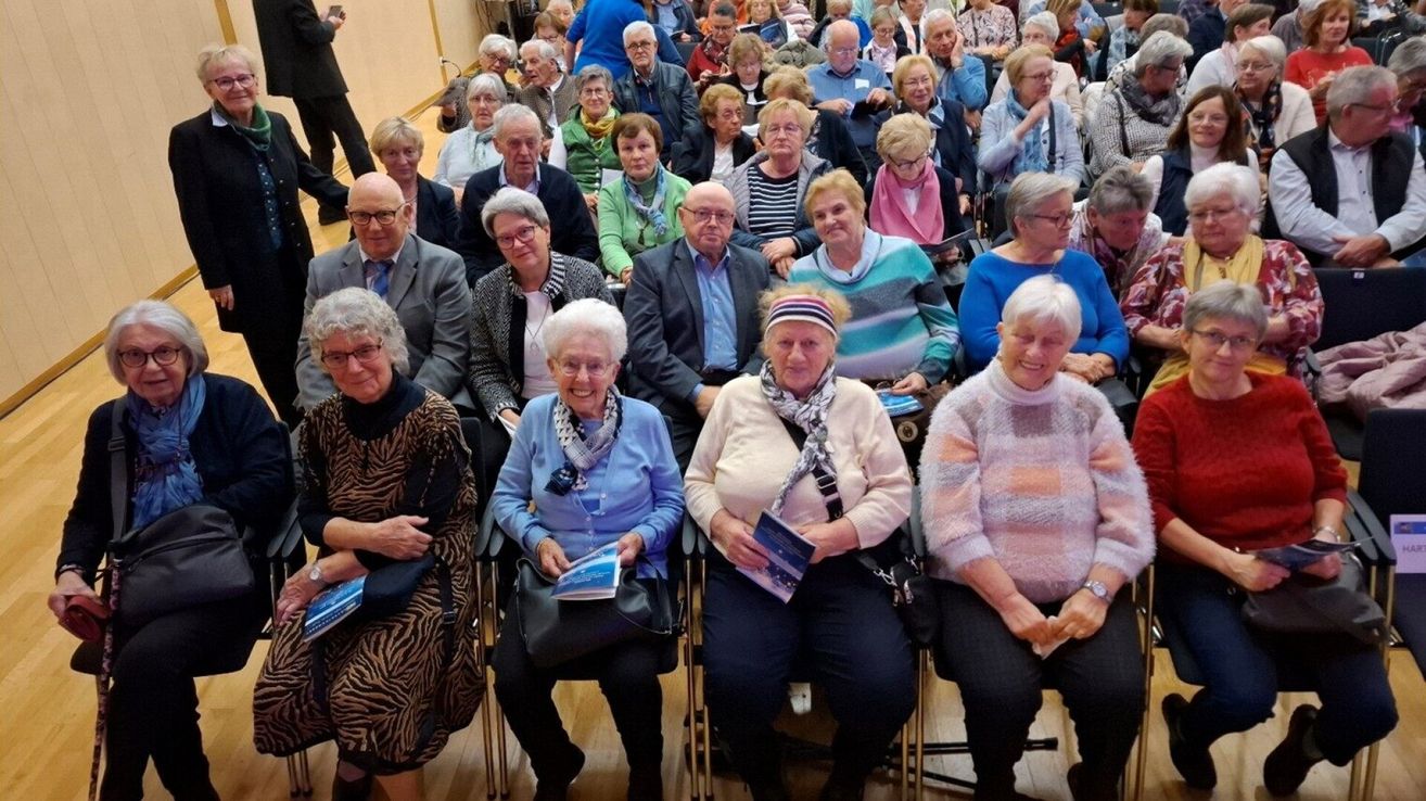Eine große Gruppe älterer Menschen sitzt gemeinsam in einem Raum, trägt Brillen, Schals und hält Bücher. Sie scheinen an einer Veranstaltung teilzunehmen.