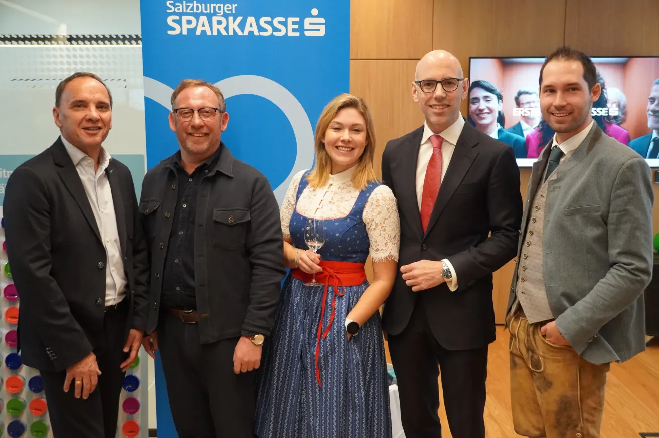 Fünf Personen stehen vor einem Salzburger Sparkasse-Banner. Eine Frau in traditioneller Kleidung hält ein Weinglas. Zwei Männer tragen Anzüge und Krawatten, die anderen lässige Kleidung.