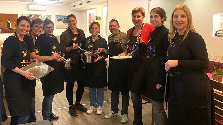 Eine Gruppe von Menschen in Schürzen und Turnschuhen posiert für ein Foto in einer Küche. Einer hält einen Kochlöffel, ein anderer einen Schneebesen und andere halten einen Teller und einen Topf.