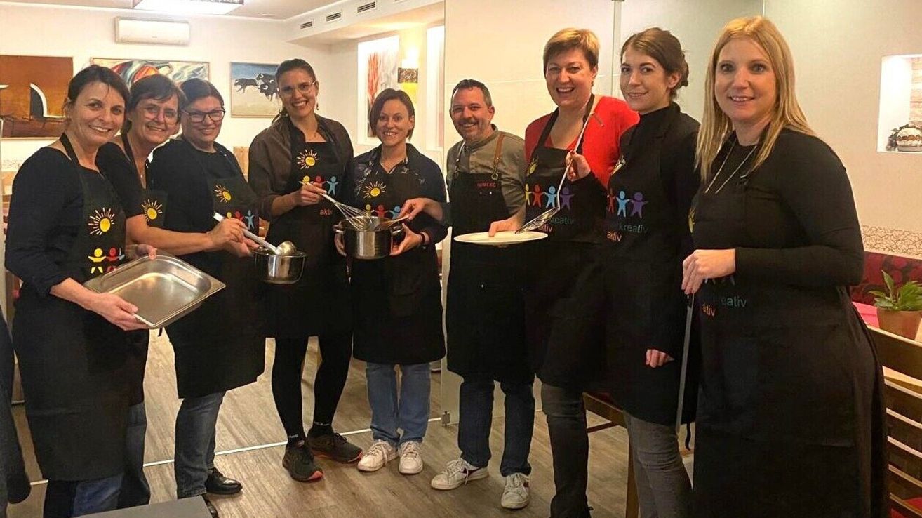 Eine Gruppe von Menschen in Schürzen und Turnschuhen posiert für ein Foto in einer Küche. Einer hält einen Kochlöffel, ein anderer einen Schneebesen und andere halten einen Teller und einen Topf.