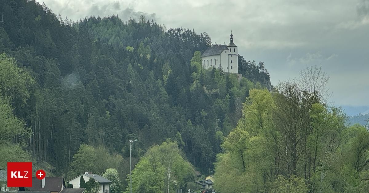 Eine Kirche mit einem Turm und mehreren Fenstern steht auf einem mit dichten grünen Bäumen bedeckten Hügel unter einem bewölkten Himmel.