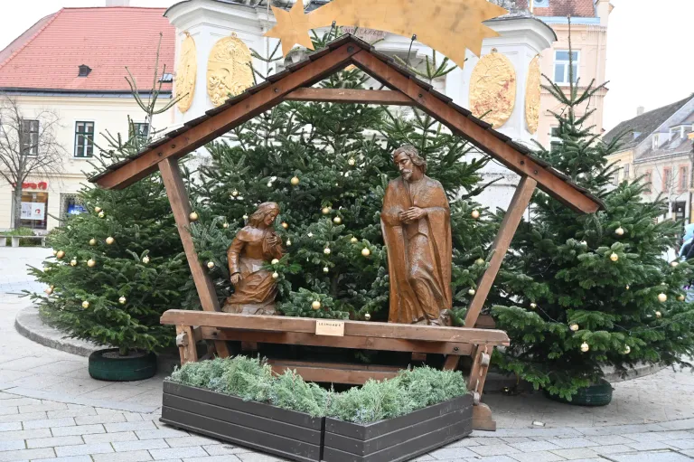 Eine Krippe mit Statuen von Jesus und Maria steht vor einem Gebäude. Sie ist von Weihnachtsbäumen und immergrünen Pflanzen umgeben. Die hölzerne Struktur ist mit einem goldenen Stern verziert.