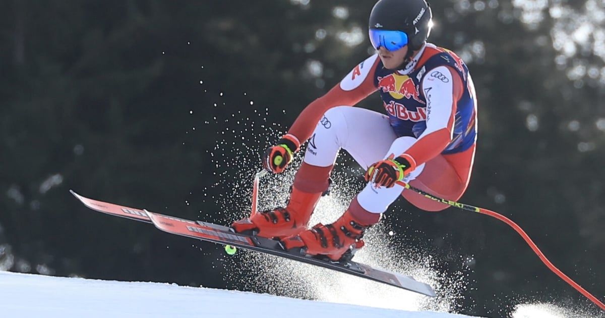 Ein Skifahrer in roter und weißer Kleidung, gesponsert von Red Bull, ist auf einem verschneiten Hang in der Luft. Der Skifahrer trägt eine Brille und Handschuhe.