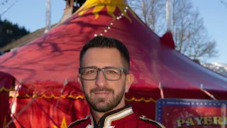 A man in a red and gold uniform stands in front of a red circus tent with the word 'GOMEZ' on top.