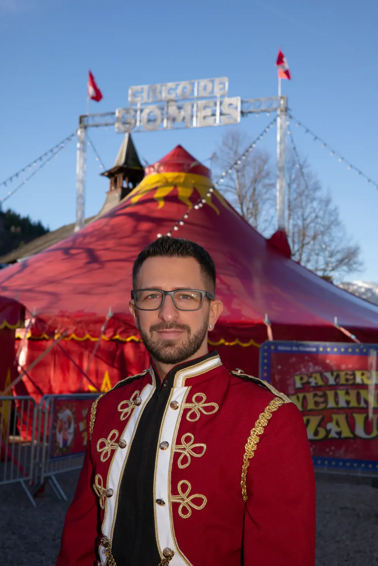 Ein Mann in einem roten und goldenen Uniform steht vor einem roten Zirkuszelt mit dem Wort 'GOMEZ' oben.