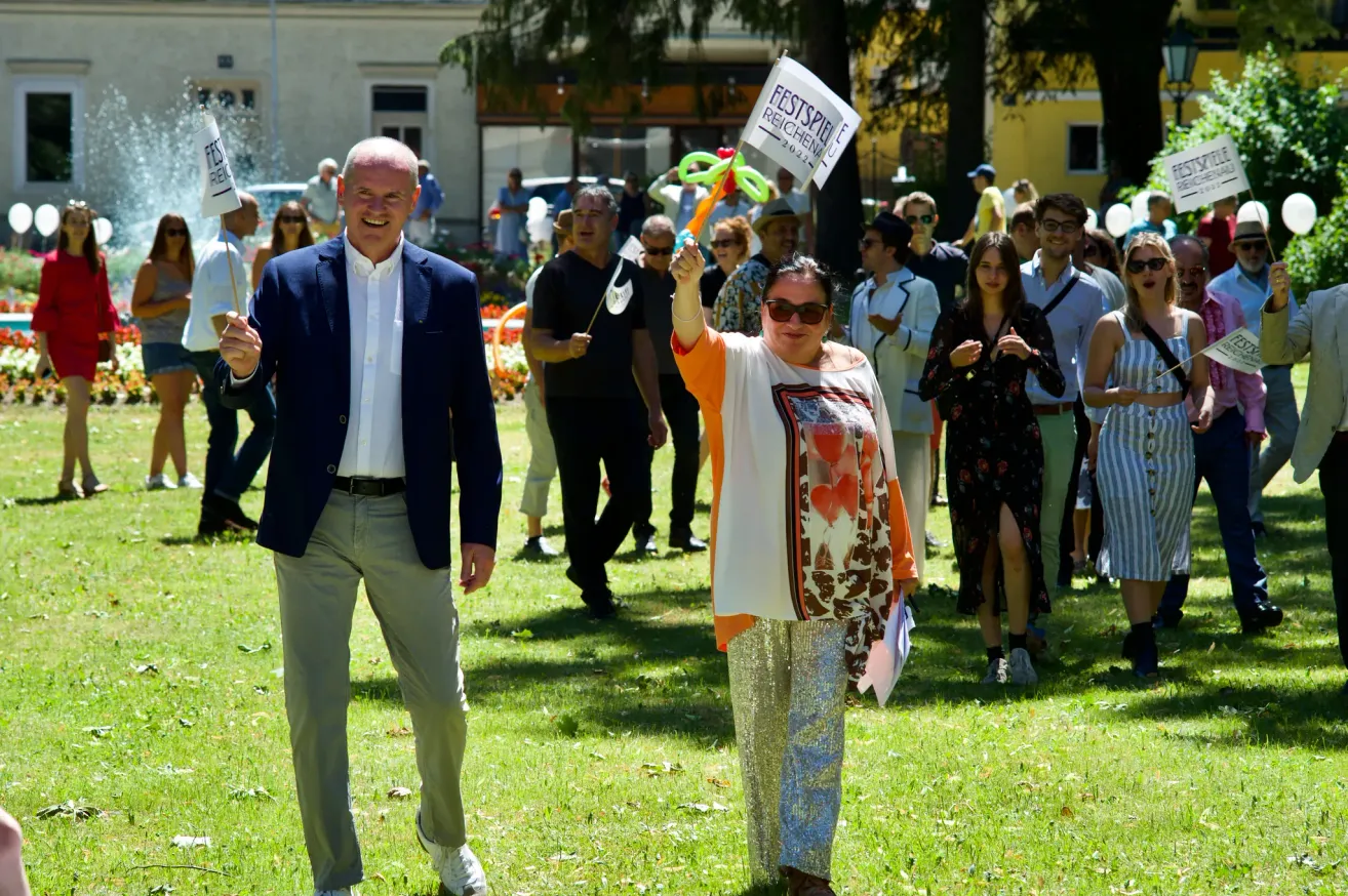 A group of people are gathered in a park. A woman holds a sign that reads 'Festspiele Reichenu'. Behind her, a man in a suit walks with a smile. Several people are standing behind them, some are clapping. In the background, there are trees and buildings.