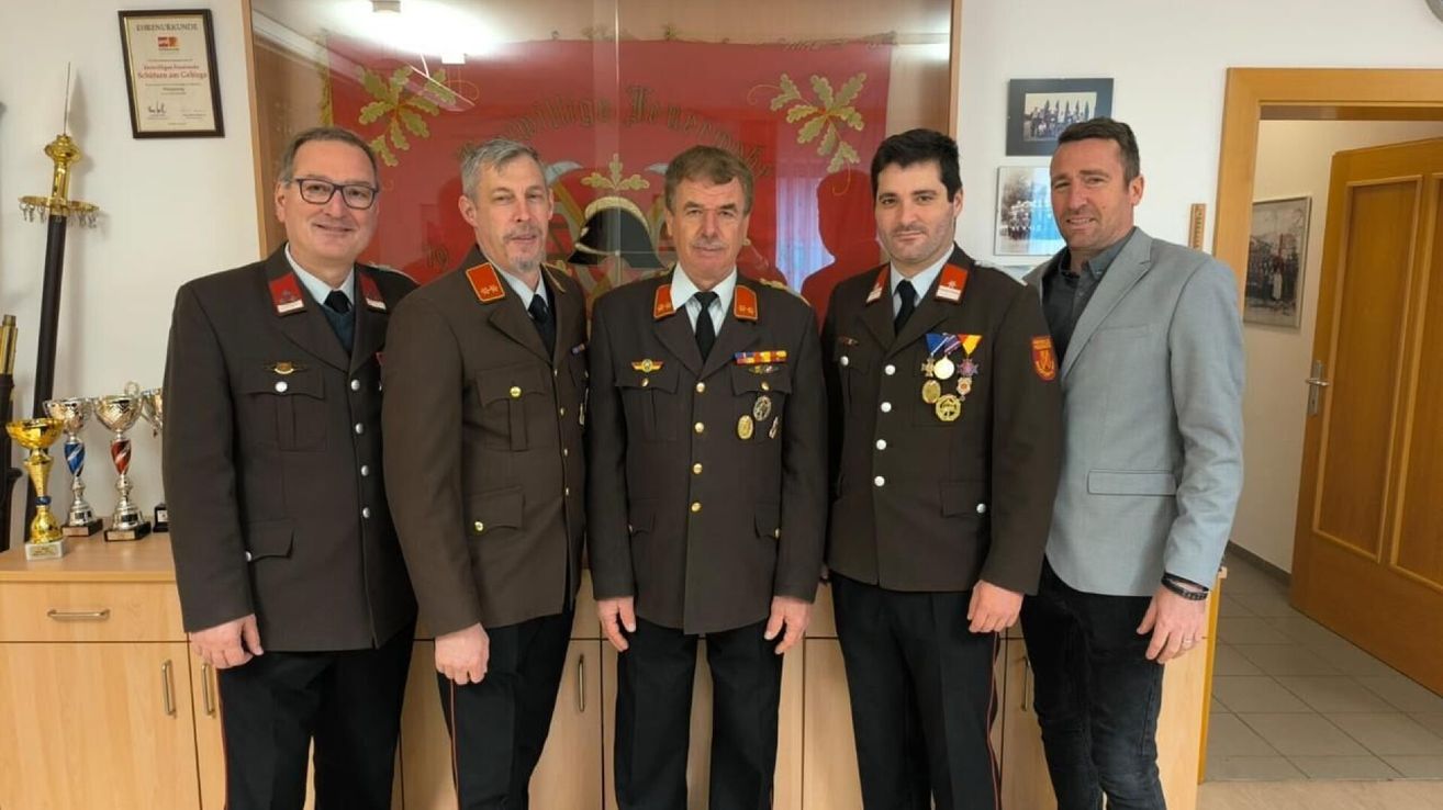 Fünf Männer stehen zusammen, gekleidet in Militäruniformen. Sie stehen vor einem roten Banner und einem Bilderrahmen.