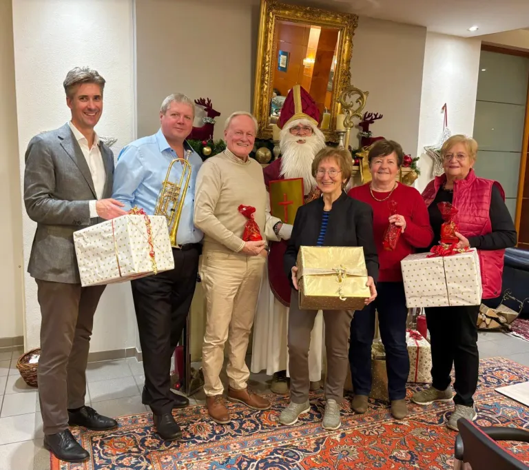 Eine Gruppe von Menschen, darunter ein älterer Mann in einer Weihnachtsmann-Kostüm, steht in einem festlichen Raum mit Geschenken und lächelt für ein Foto.